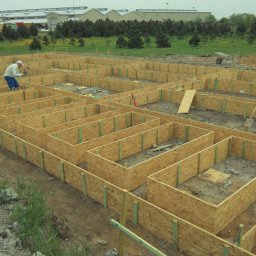 Mac Bud Maciej Birecki - Szalunek fundamentów domu jednorodzinnego z płyty OSB, wzmocniony pionowymi listwami, pracownik wbija gwoździe, w tle teren budowy i budynki gospodarcze.