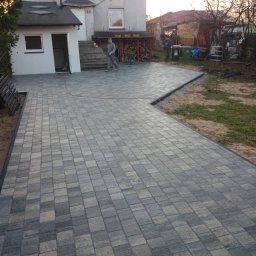 Mac Bud Maciej Birecki - Ułożona szara kostka brukowa na podjeździe przed domem jednorodzinnym z elementami ogrodu w tle, widoczni dwaj mężczyźni i pies.