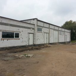 Mac Bud Maciej Birecki - Budynek w trakcie budowy z białych bloczków, widoczne otwory okienne i drzwiowe, teren wokół budynku ziemisty i nierówny, pochmurne niebo.