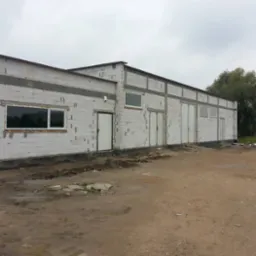 Budynek w trakcie budowy z białych bloczków, widoczne otwory okienne i drzwiowe, teren wokół budynku ziemisty i nierówny, pochmurne niebo.