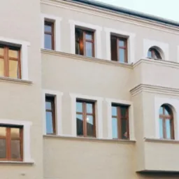 Fragment elewacji budynku z oknami o drewnianej stolarce, wykończonej w jasnym odcieniu z dekoracyjnymi obramowaniami okiennymi i gzymsami.