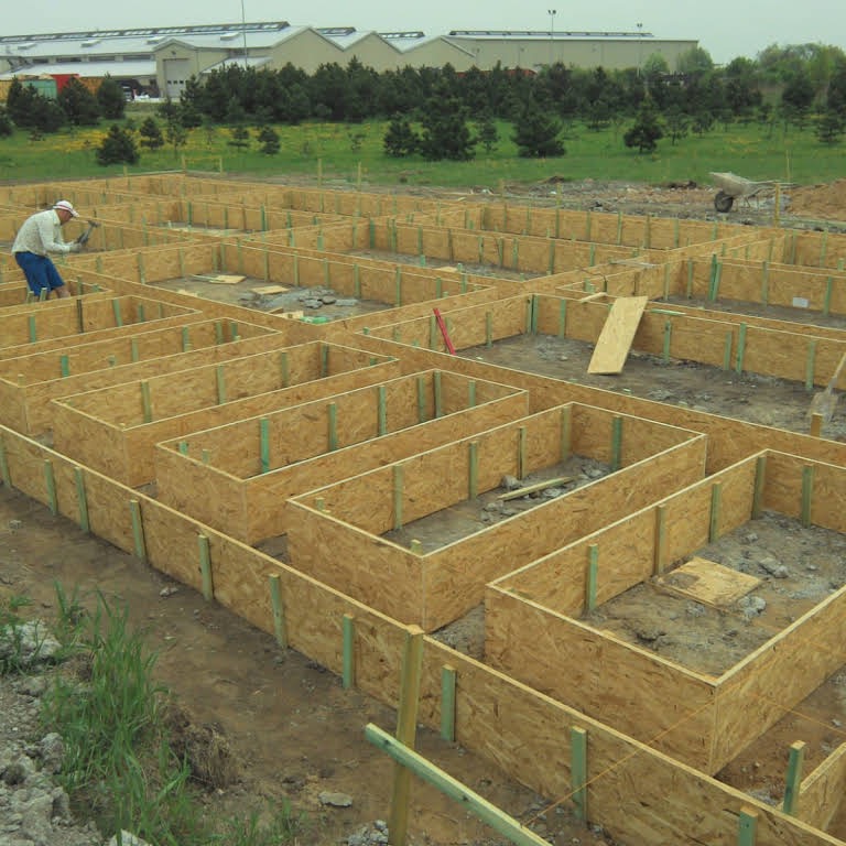 Szalunek fundamentów domu jednorodzinnego z płyty OSB, wzmocniony pionowymi listwami, pracownik wbija gwoździe, w tle teren budowy i budynki gospodarcze.