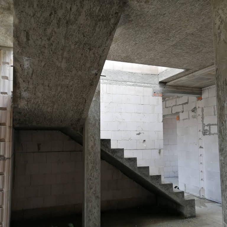 Surowe wnętrze domu w trakcie budowy, widoczne betonowe schody, ściany z białych bloczków i surowy betonowy strop.