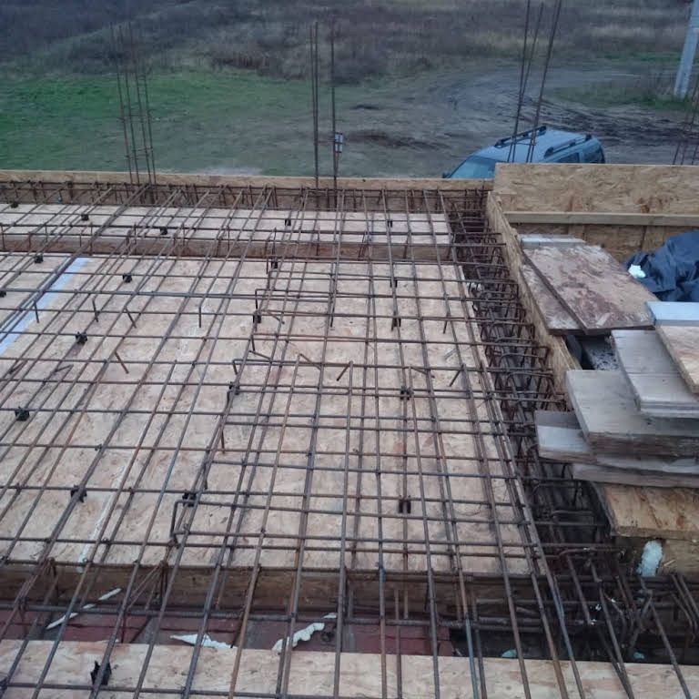 Zbrojenie stropu na deskowaniu z płyty OSB, widoczne wystające pręty pionowe, w tle krajobraz polny i zaparkowany samochód.