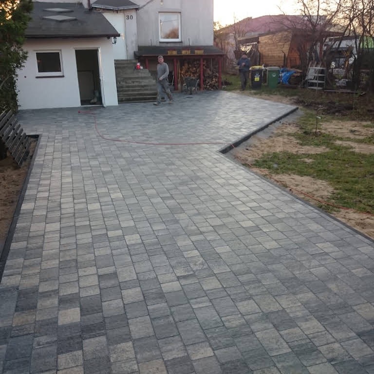 Ułożona szara kostka brukowa na podjeździe przed domem jednorodzinnym z elementami ogrodu w tle, widoczni dwaj mężczyźni i pies.