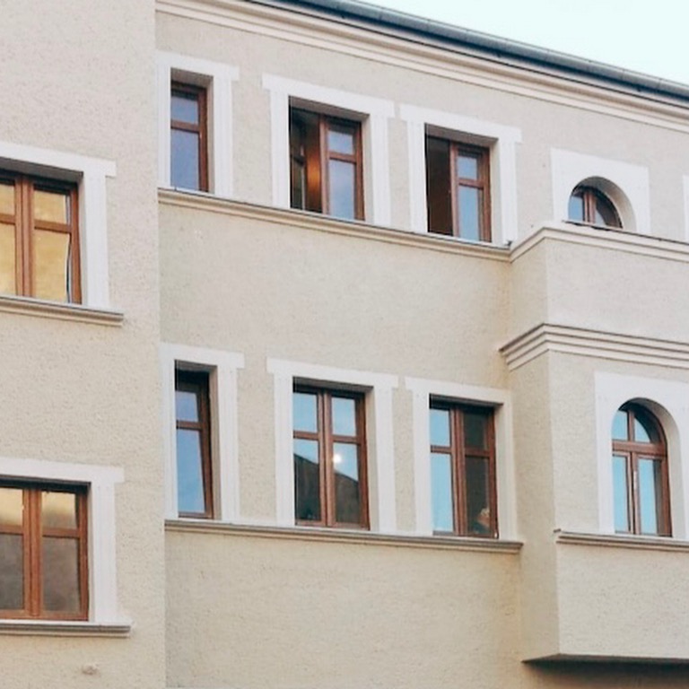 Fragment elewacji budynku z oknami o drewnianej stolarce, wykończonej w jasnym odcieniu z dekoracyjnymi obramowaniami okiennymi i gzymsami.