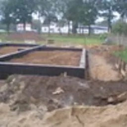 Zabezpieczone fundamenty budynku, pokryte czarną izolacją, wypełnione piaskiem, widoczne z poziomu gruntu, w tle domy jednorodzinne i drzewa.