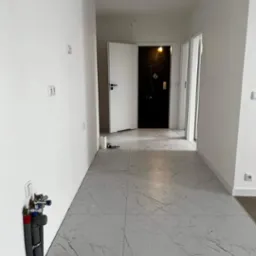 Wnętrze korytarza w trakcie remontu z szarymi płytkami imitującymi marmur na podłodze, widoczne instalacje wodno-kanalizacyjne przy ścianie, białe ściany z zamontowanymi gniazdkami elektrycznymi...