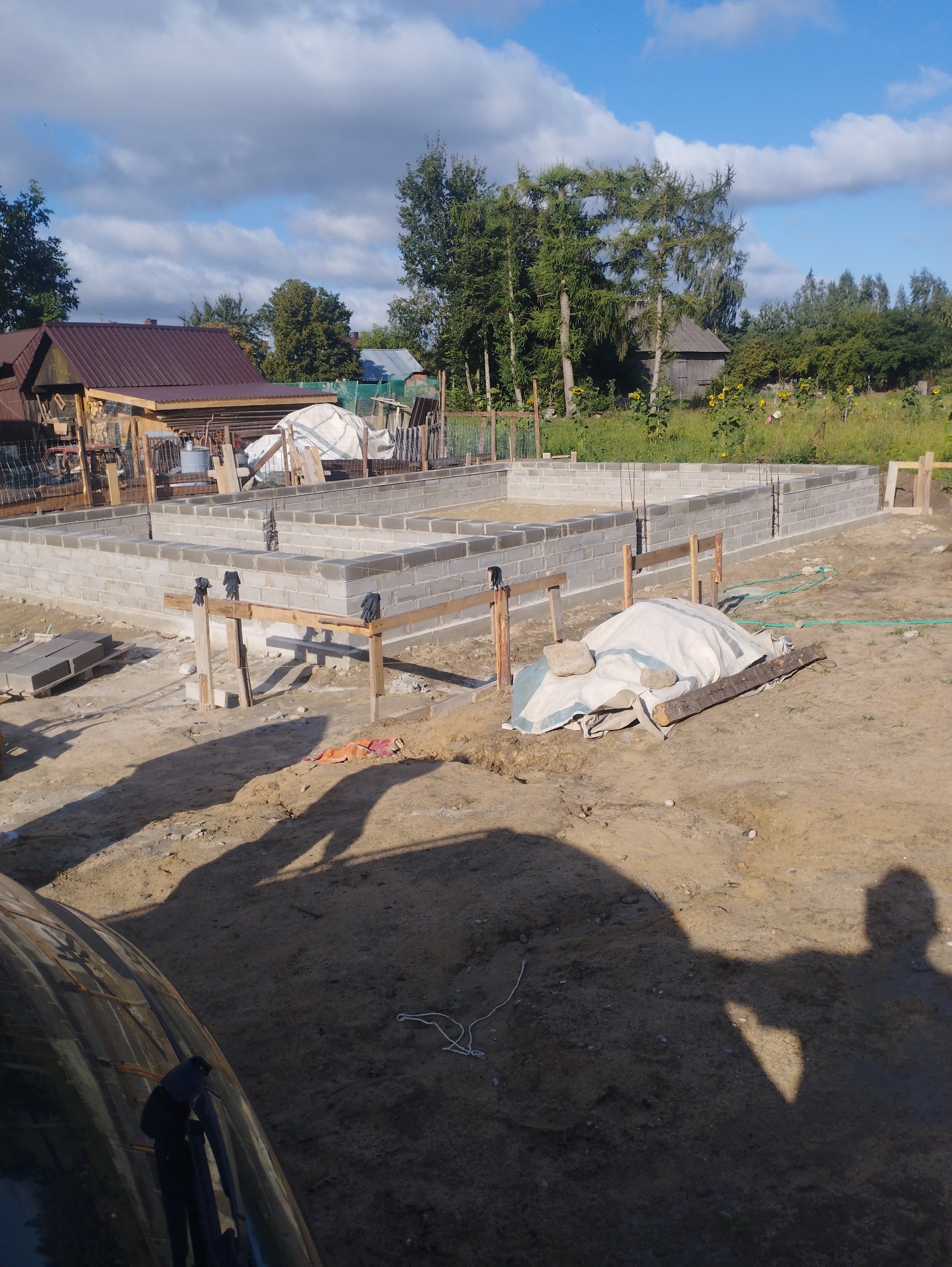 Wznoszenie fundamentów domu jednorodzinnego z bloczków betonowych, widoczne zbrojenia pionowe, drewniane podpory i zakryte materiały budowlane na placu budowy w słoneczny dzień.