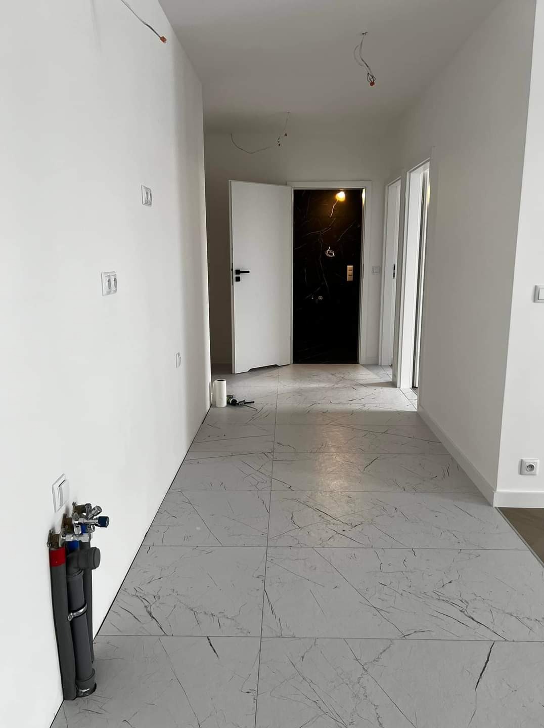 Wnętrze korytarza w trakcie remontu z szarymi płytkami imitującymi marmur na podłodze, widoczne instalacje wodno-kanalizacyjne przy ścianie, białe ściany z zamontowanymi gniazdkami elektrycznymi...