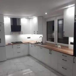 Biała, nowoczesna kuchnia z drewnianym blatem, czarnymi uchwytami i marmurową podłogą, oświetlona listwami LED pod szafkami wiszącymi.