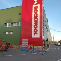 Montaż elewacji z wełny mineralnej Rockwool na budynku przemysłowym, widoczny podnośnik koszowy i pracownik w kasku.