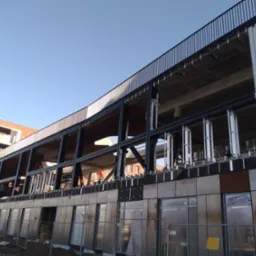 Wielopiętrowy budynek w trakcie budowy, z widoczną stalową konstrukcją i częściowo zamontowaną elewacją z metalowych paneli. Widoczne okna i barierki na górnej kondygnacji.