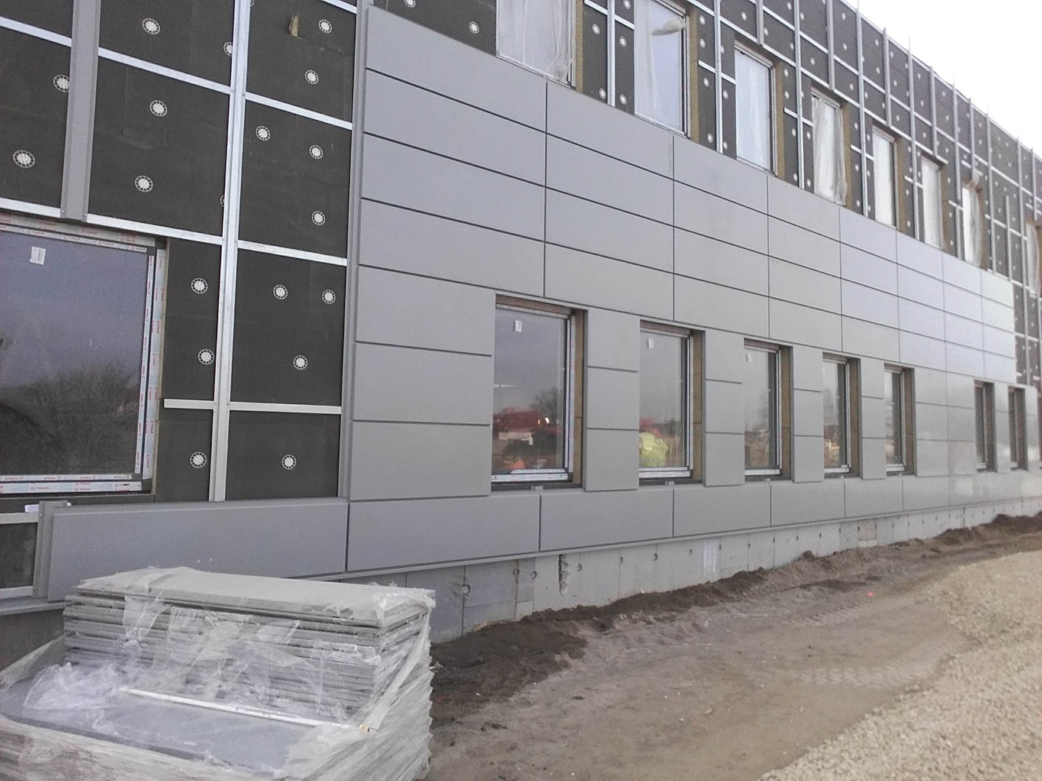 Montaż elewacji z szarych paneli na budynku, widoczne warstwy ocieplenia i okna, na dole stos paneli w folii.