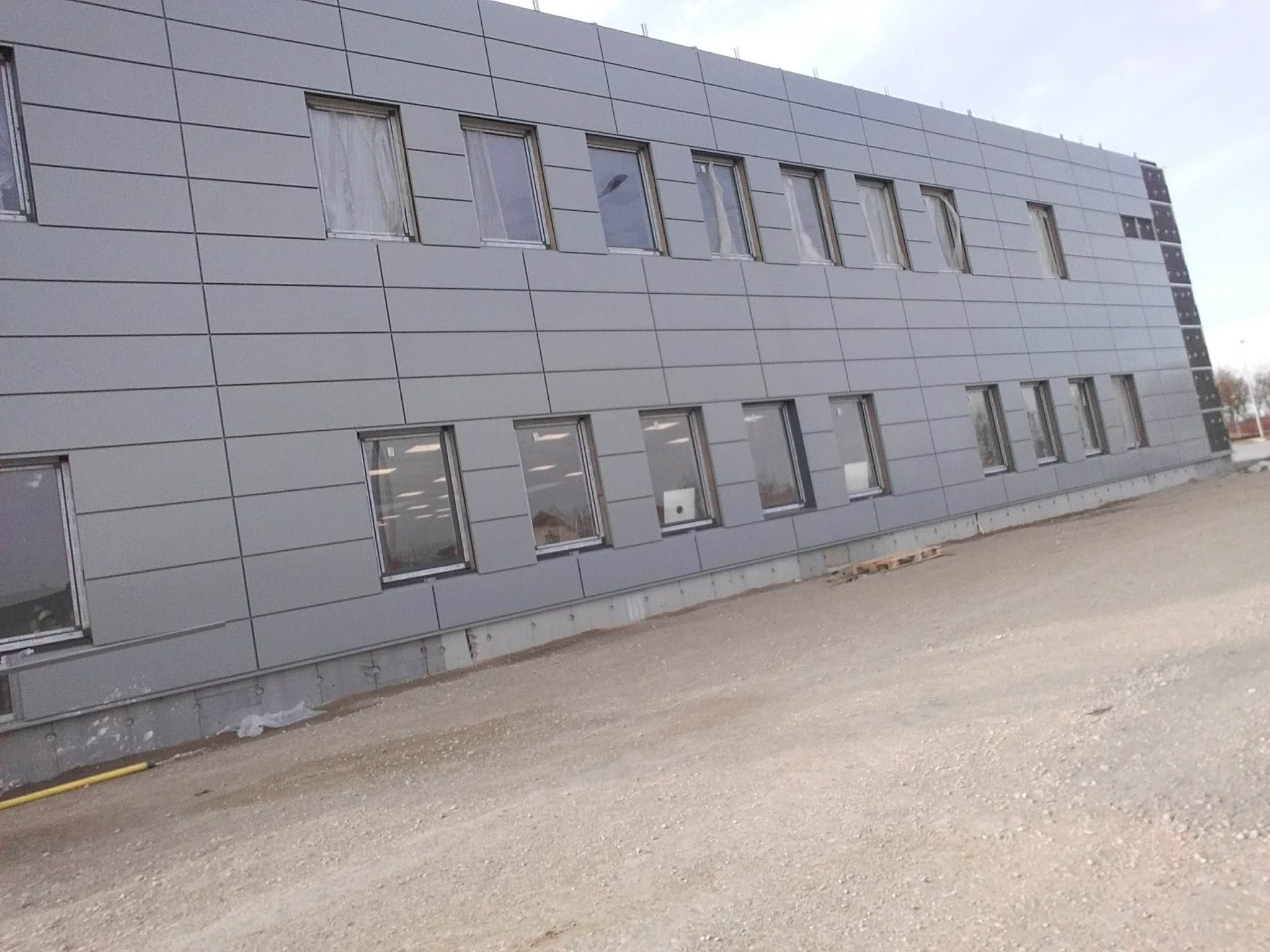 Szara elewacja budynku biurowego z panelami kompozytowymi, widoczne okna z zasłonami i teren budowy z gruzem.