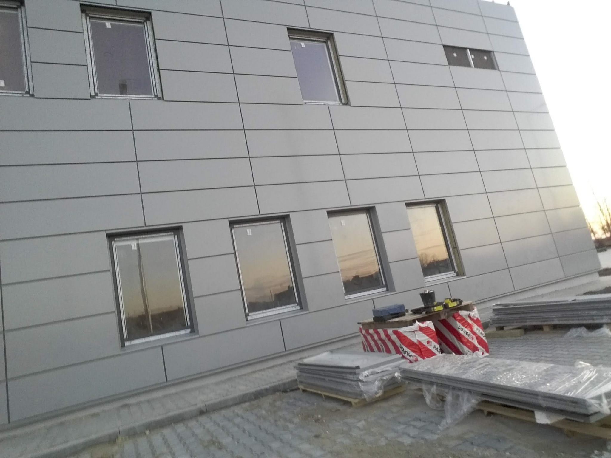 Montaż elewacji z paneli kompozytowych na budynku, widoczne okna z odbijającym się zachodem słońca, palety z materiałami budowlanymi na pierwszym planie.