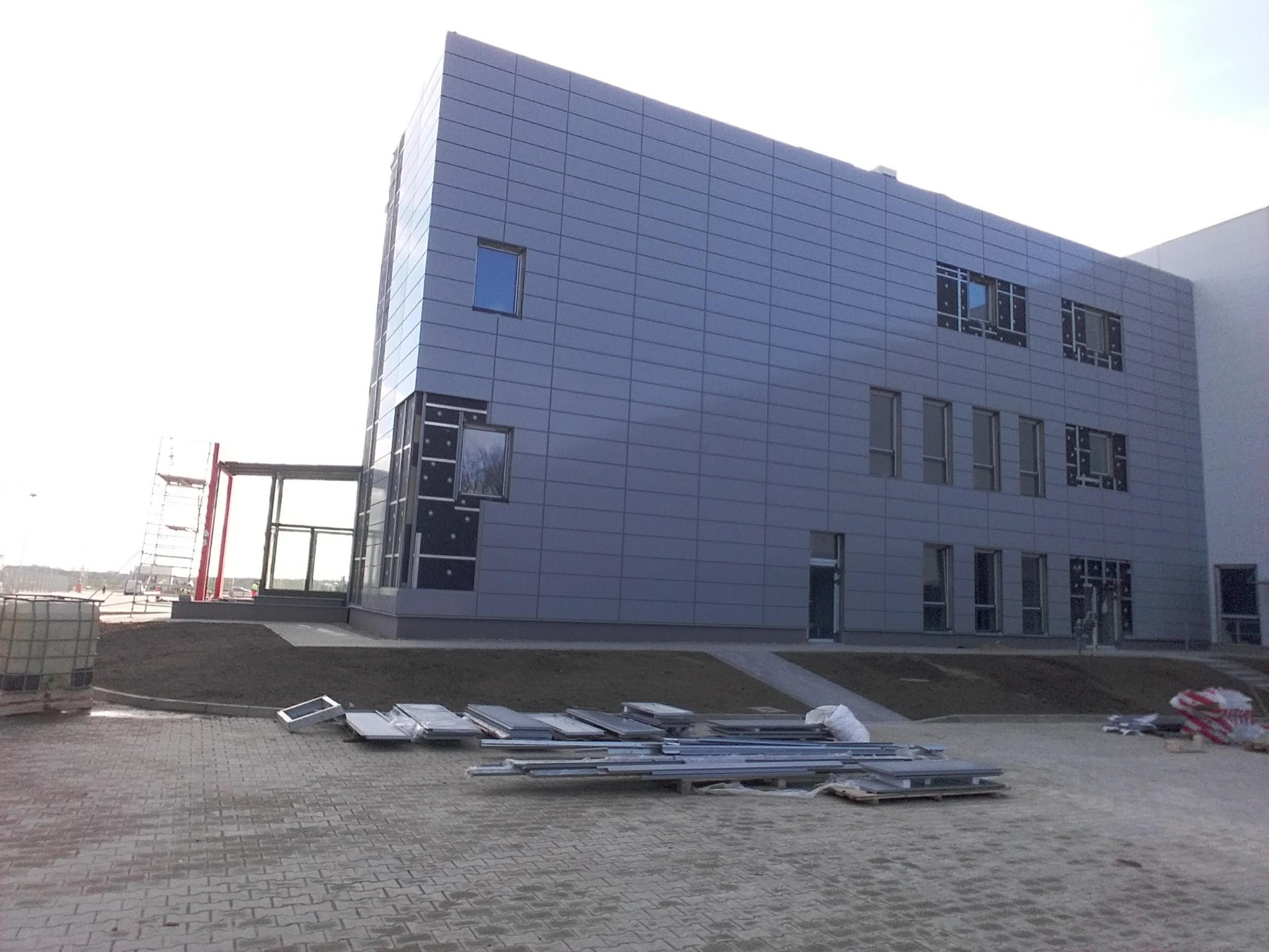 Montaż szarej elewacji panelowej na budynku przemysłowym, widoczne panele elewacyjne na ziemi, częściowo zamontowane okna, rusztowanie w tle.