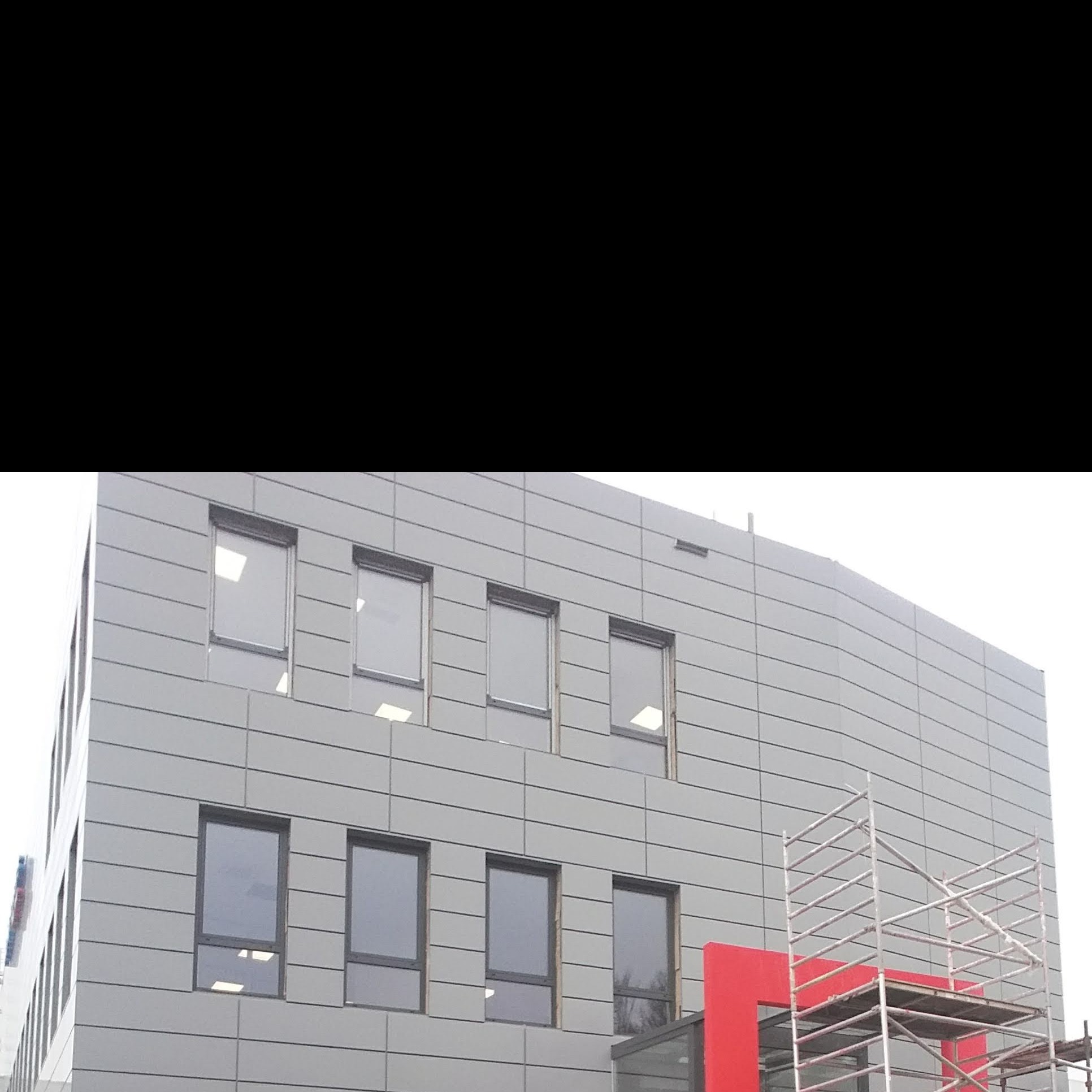 Nowoczesny budynek biurowy z szarą elewacją panelową, widoczne rusztowania i czerwone elementy architektoniczne.
