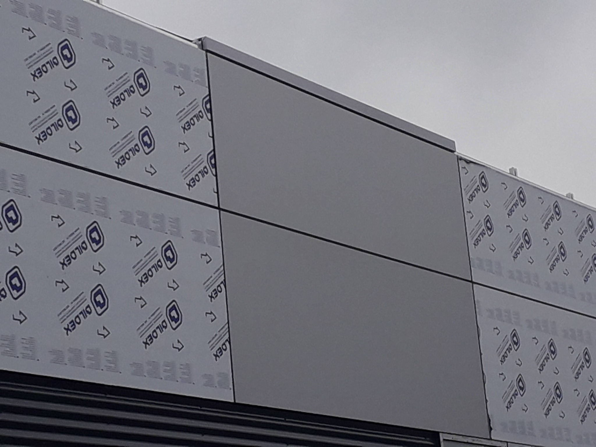 Fragment elewacji budynku z paneli kompozytowych, częściowo pokrytych folią ochronną z nadrukowanym logo producenta DILDEX, widoczne łączenia paneli i metalowe profile.