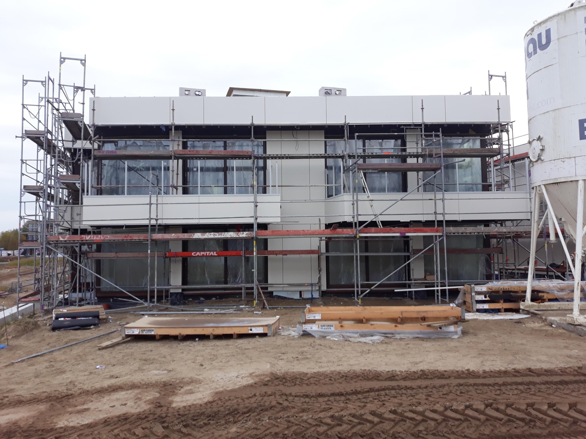 Budynek biurowy w trakcie budowy, z widocznymi rusztowaniami, elementami elewacji i zbiornikiem na zaprawę na placu budowy. Widoczne palety z materiałami budowlanymi.