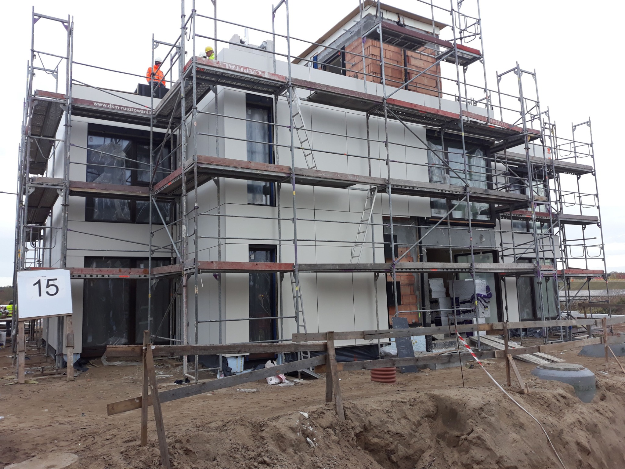 Nowoczesny dom w trakcie budowy z białymi panelami elewacyjnymi, otoczony rusztowaniami i z numerem posesji '15' na tablicy.