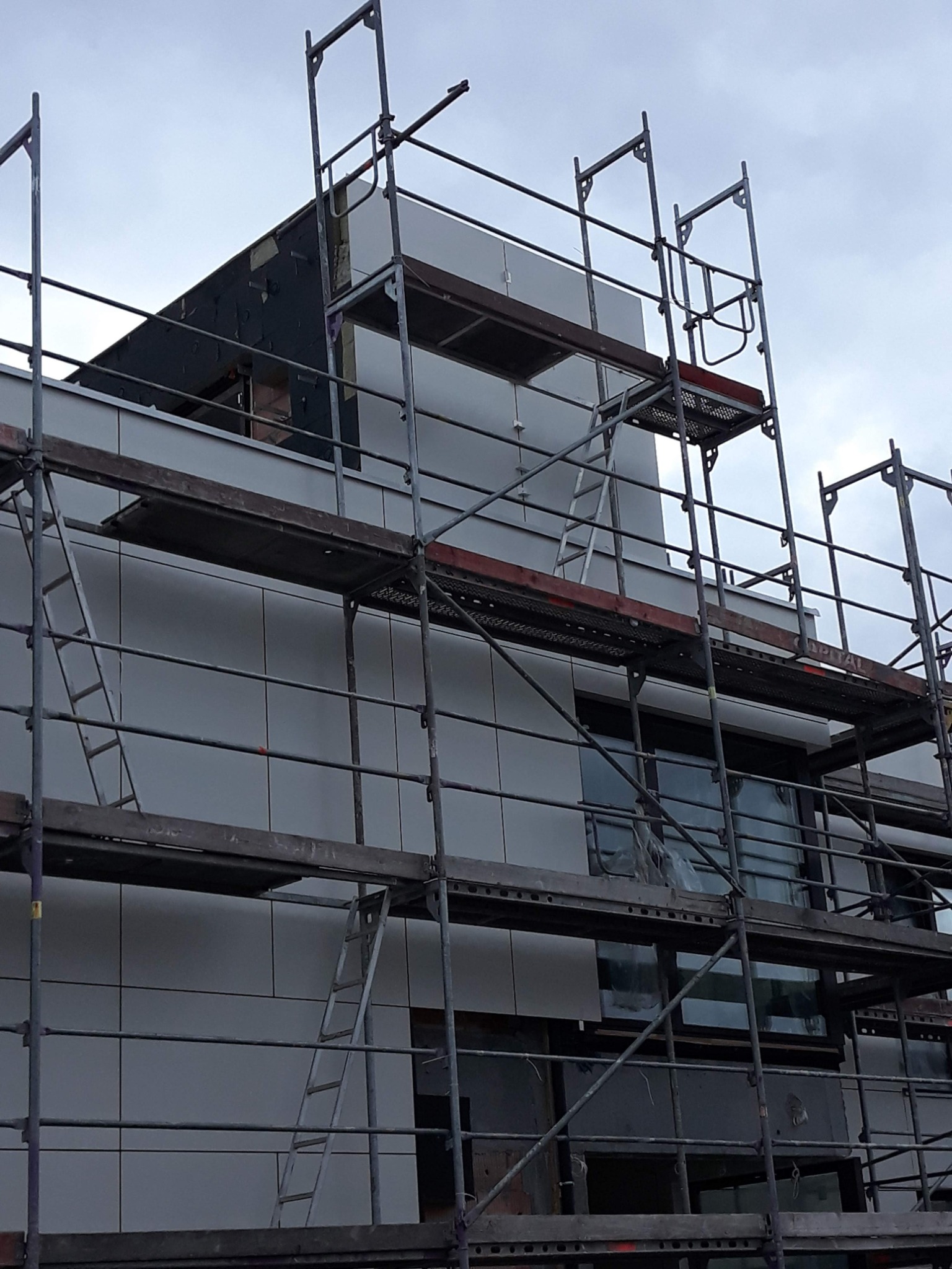 Budynek w trakcie prac elewacyjnych, widoczne rusztowania i częściowo ocieplona ściana z szarymi panelami, fragment dachu z widocznym ociepleniem.