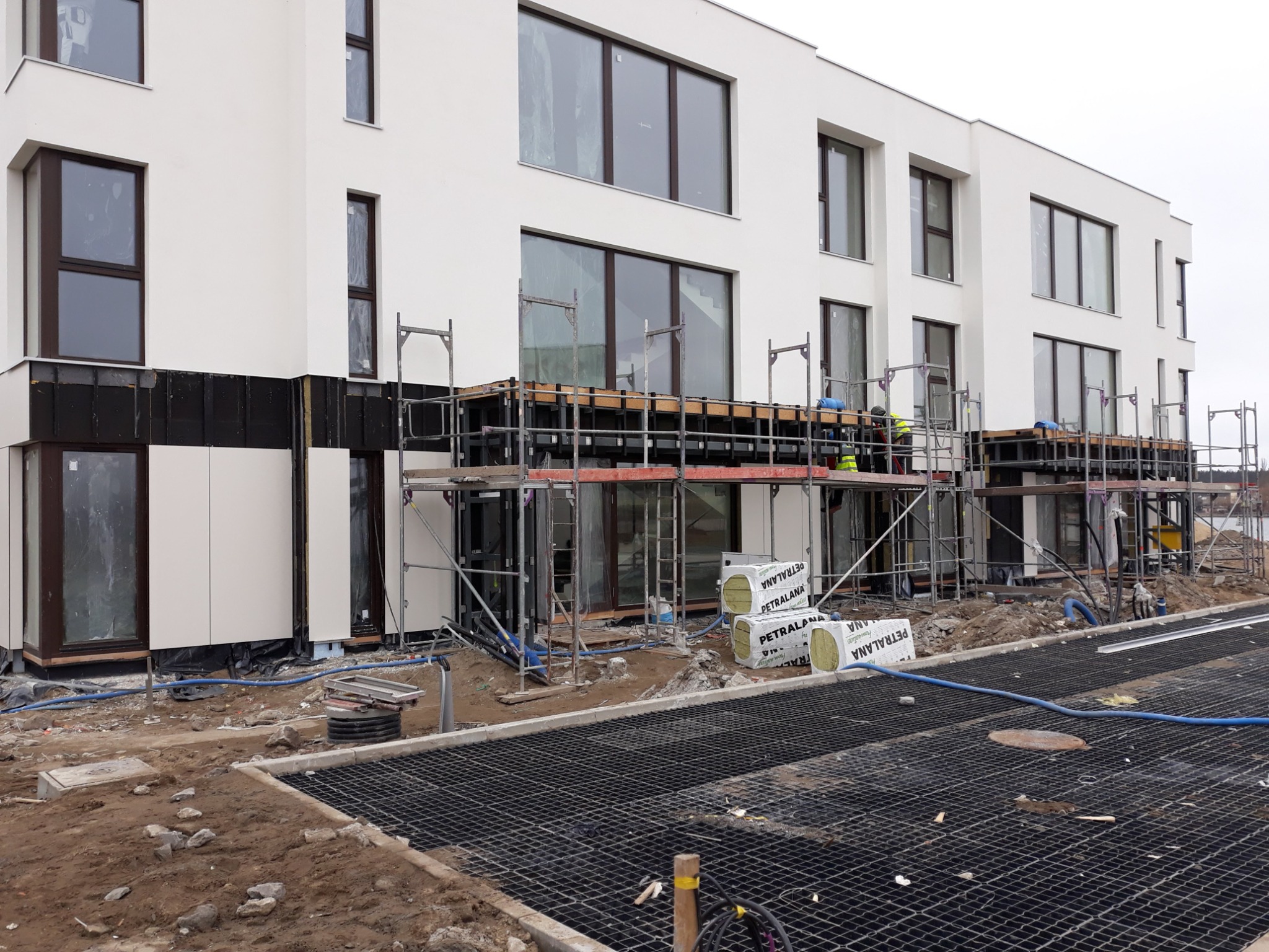 Budynek w trakcie prac elewacyjnych: widoczne rusztowania, częściowo zamontowana elewacja, oraz materiały izolacyjne Petralana na placu budowy. Część elewacji w kolorze białym, część w ciemnym...