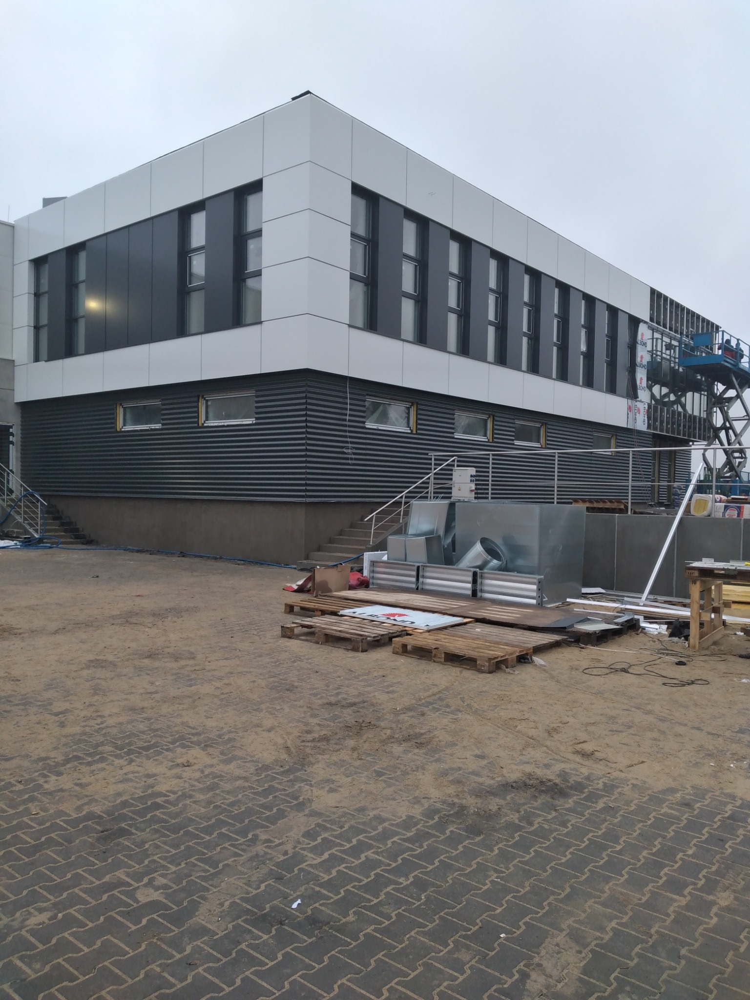 Nowoczesny budynek biurowy z szarą, poziomą elewacją i białymi panelami na górze, widoczny w trakcie prac wykończeniowych. Na pierwszym planie palety i elementy wentylacji, w tle podnośnik koszowy.
