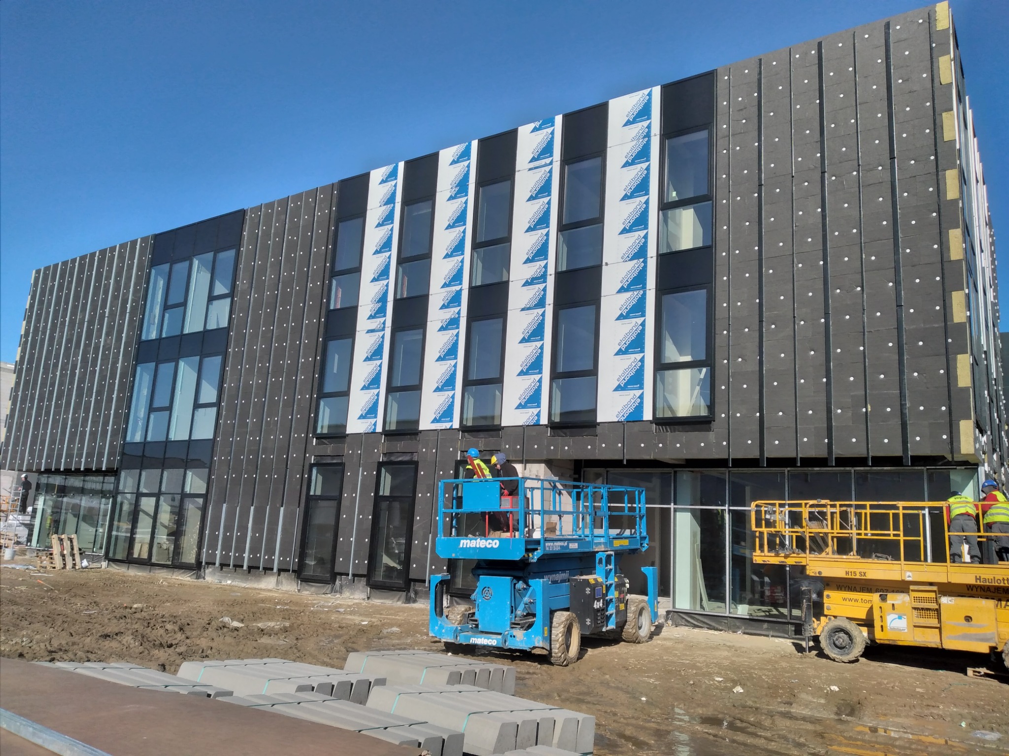 Dwupiętrowy budynek komercyjny w trakcie prac elewacyjnych, widoczne rusztowania i podnośniki koszowe, częściowo ocieplona ściana z szarymi panelami i białymi kołkami, fragmenty elewacji...