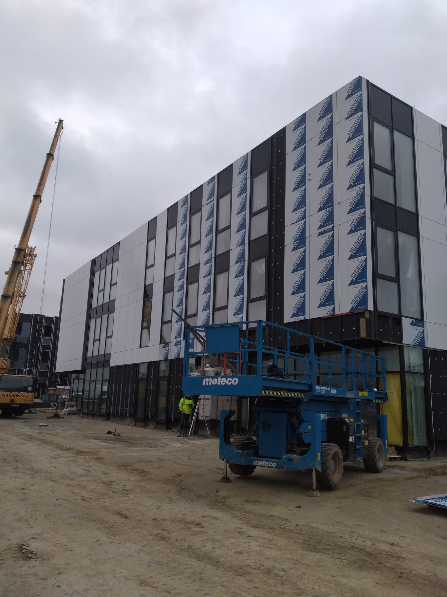 Budynek biurowy w trakcie montażu elewacji z płyt kompozytowych, widoczny dźwig i podnośnik koszowy na placu budowy, pochmurne niebo.