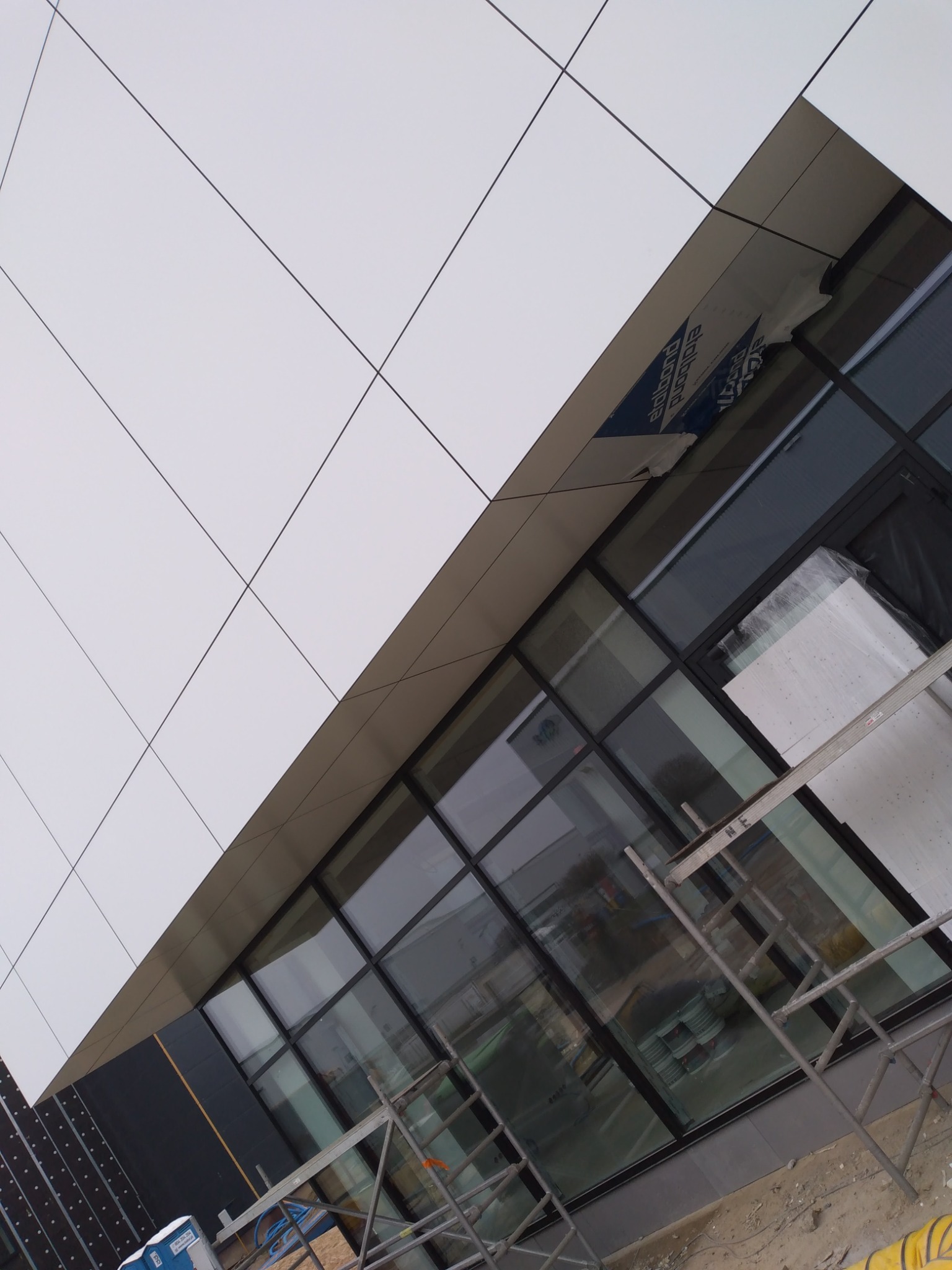 Nowoczesna elewacja budynku biurowego w trakcie montażu, widoczne białe panele elewacyjne z czarnymi fugami, okna w czarnych ramach, rusztowanie oraz materiały budowlane.