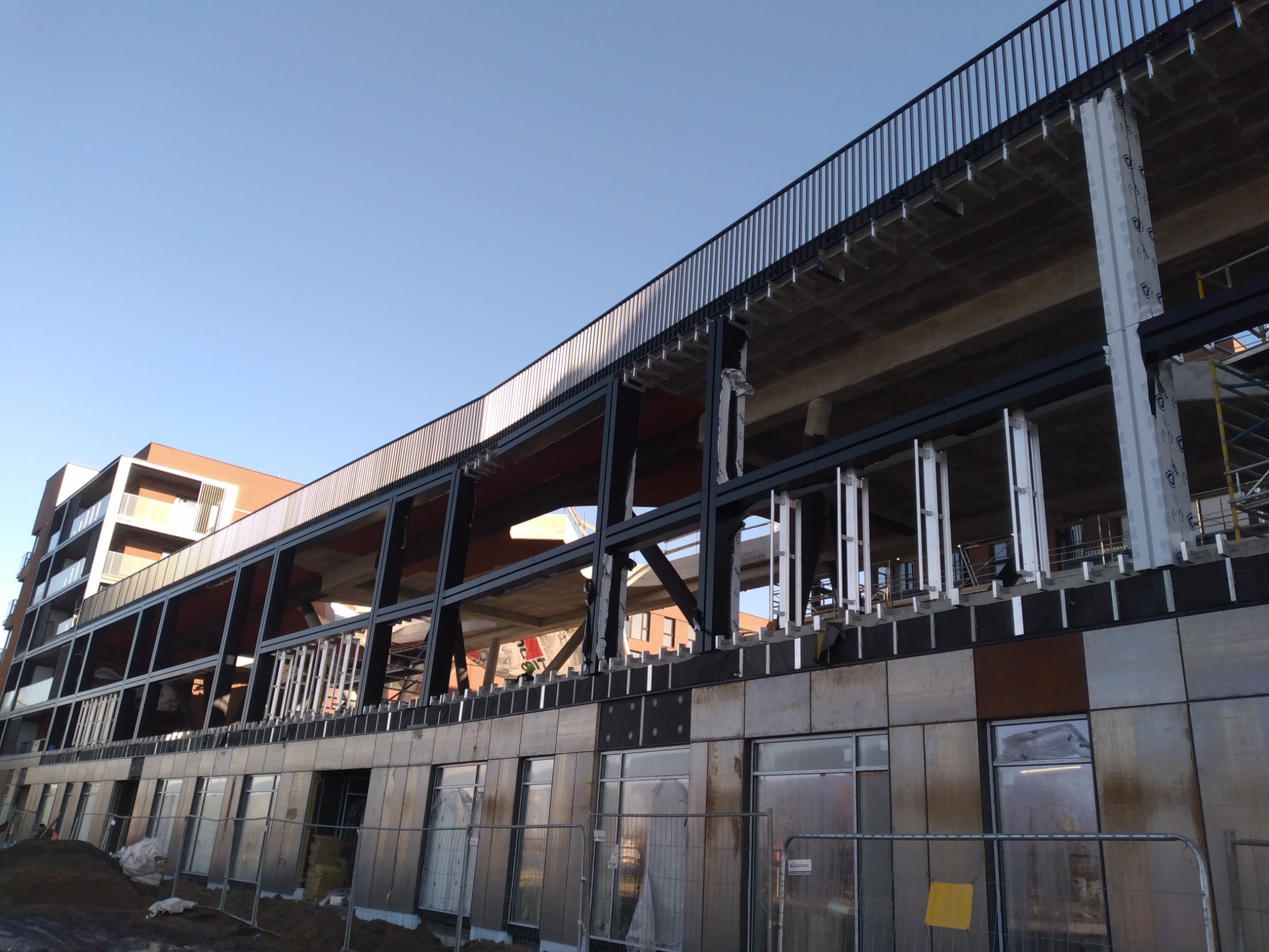 Wielopiętrowy budynek w trakcie budowy, z widoczną stalową konstrukcją i częściowo zamontowaną elewacją z metalowych paneli. Widoczne okna i barierki na górnej kondygnacji.