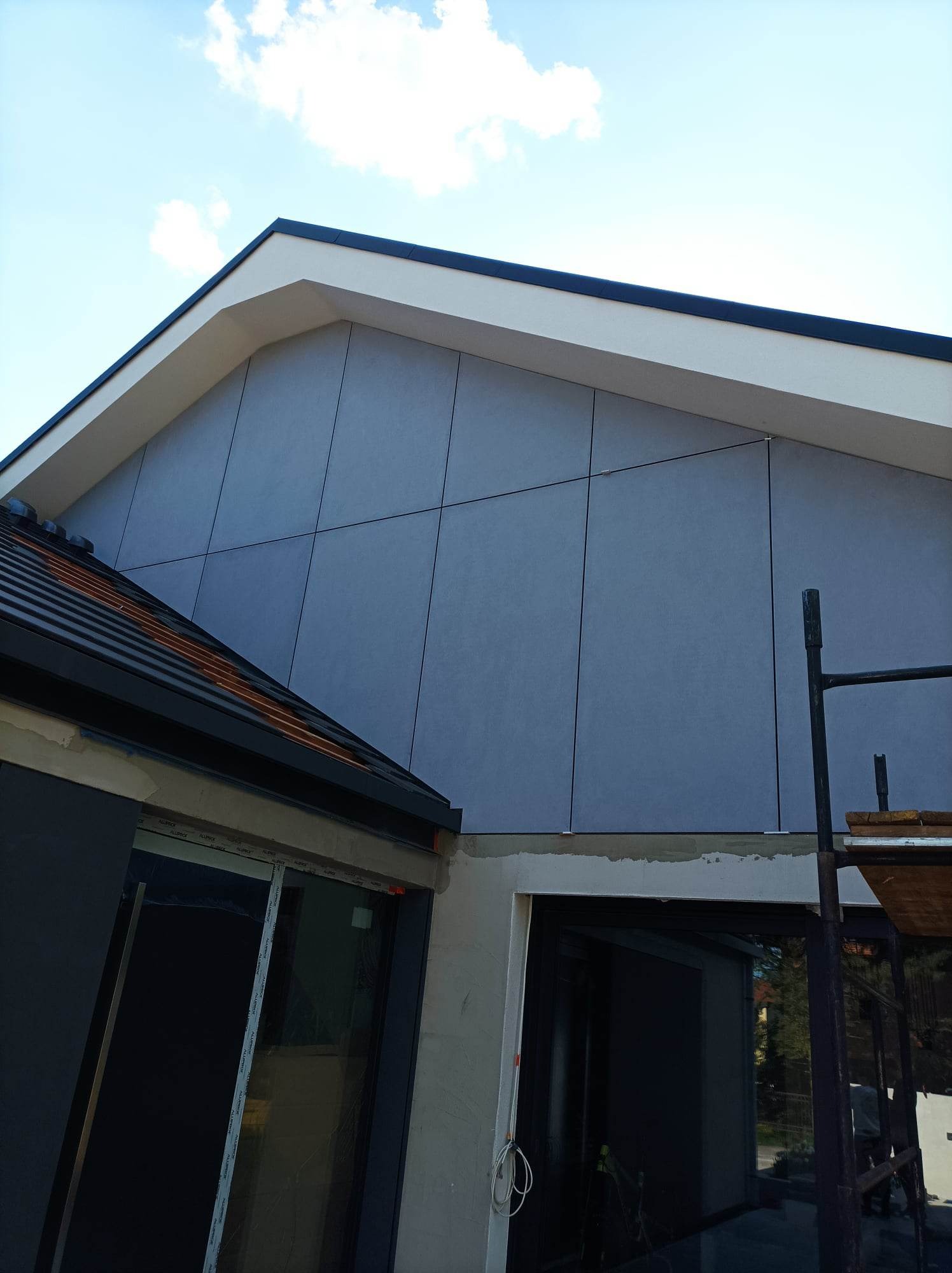 Fragment nowoczesnego domu z szarą elewacją panelową, widoczne rusztowanie i fragment dachu z czarną blachodachówką, błękitne niebo w tle.