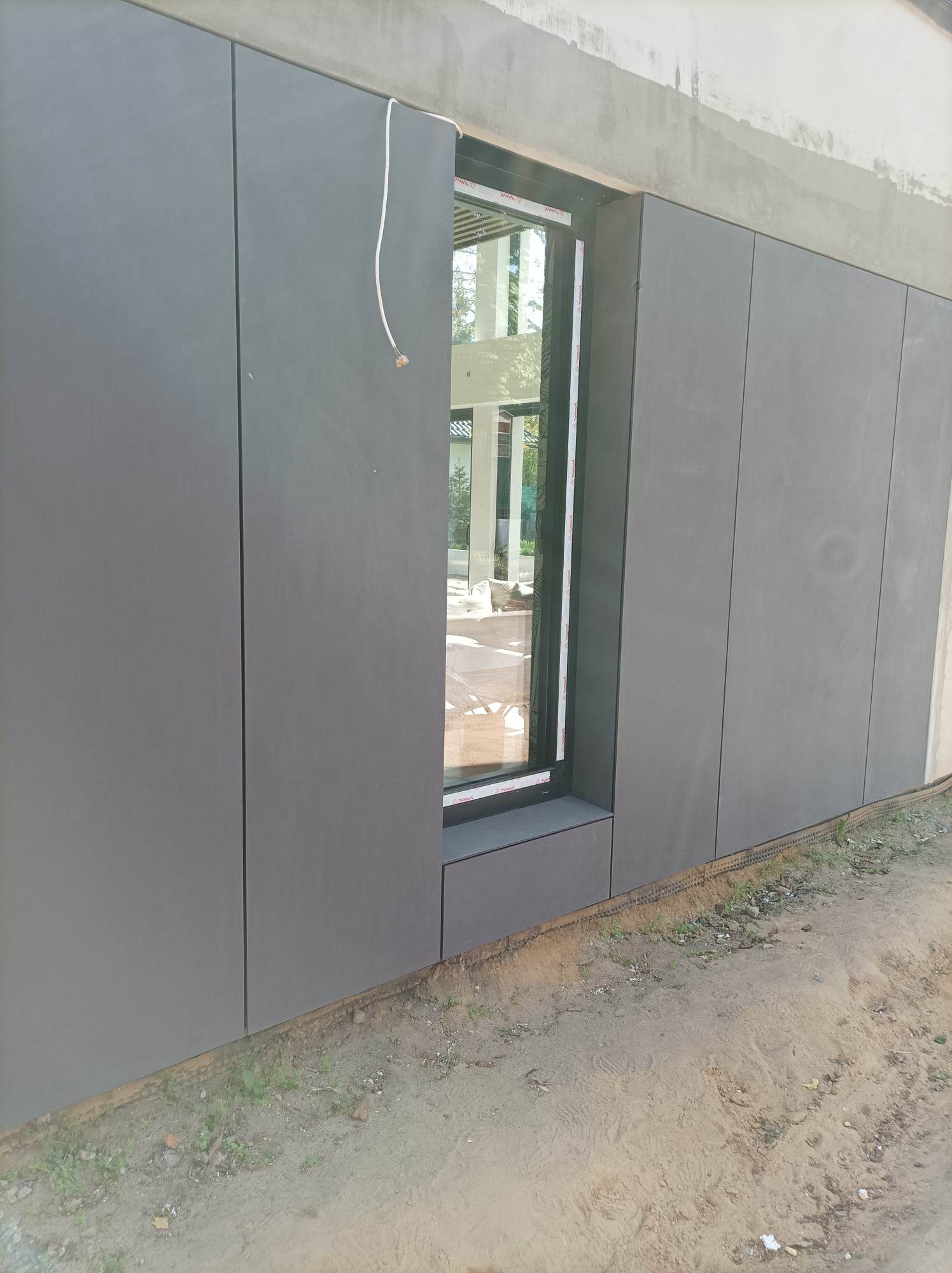 Szare panele elewacyjne zamontowane wokół okna na budynku w stanie surowym, widoczne kable elektryczne wystające z górnej krawędzi panelu.