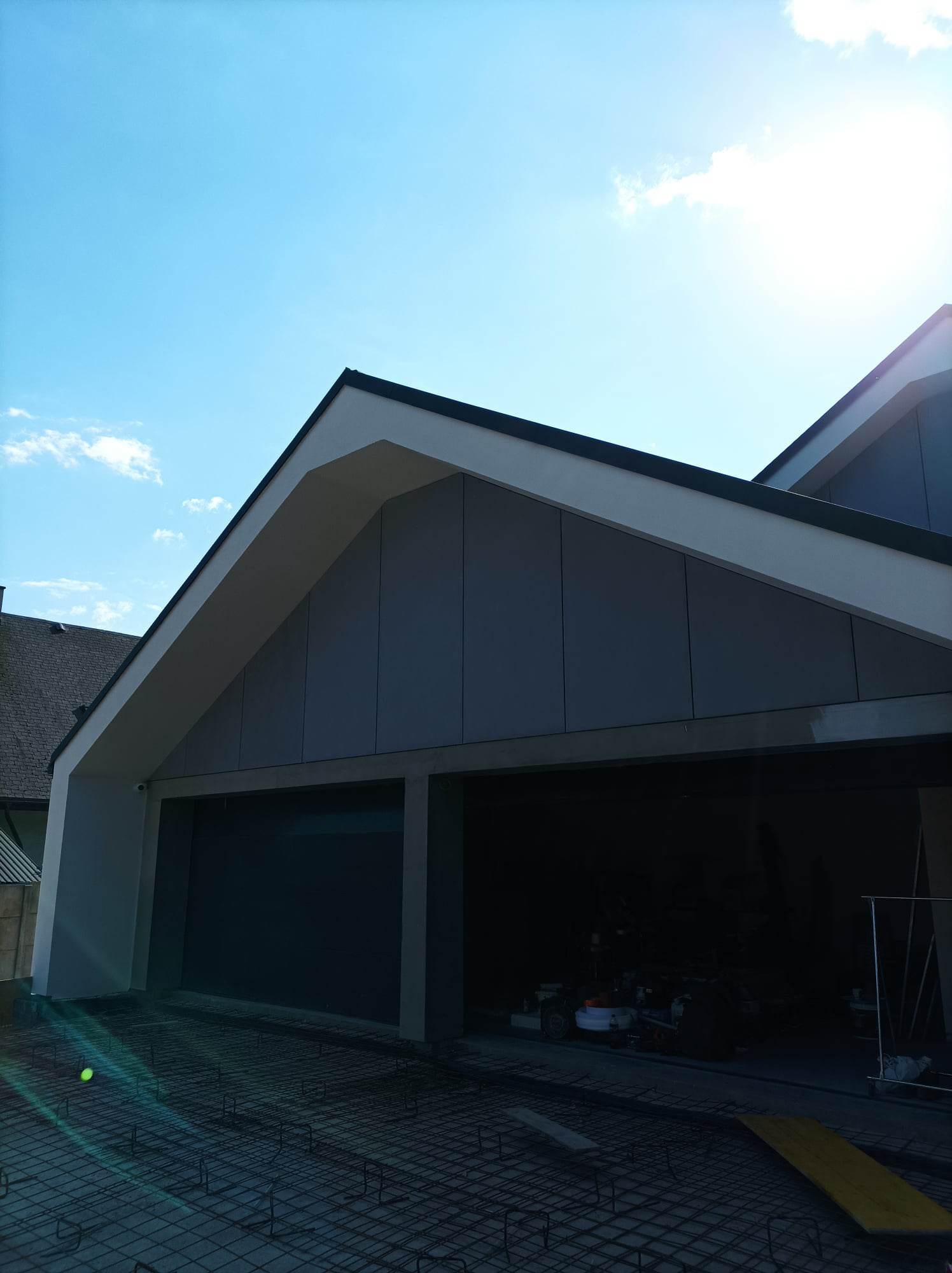 Nowoczesny dom z garażem w trakcie budowy, widoczne zbrojenie pod wylewkę, elewacja wykończona panelami w odcieniach szarości, jasne niebo.