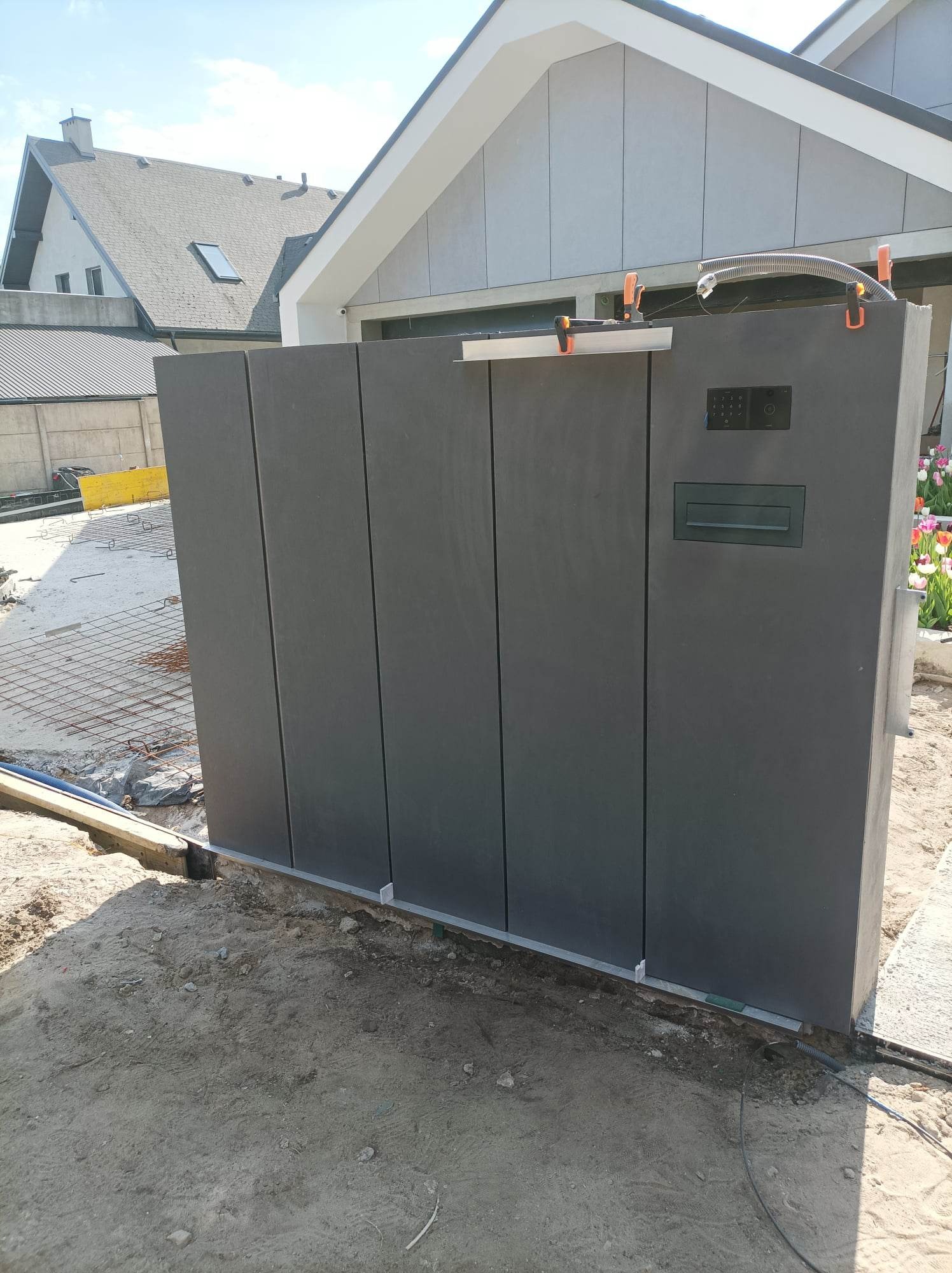 Nowoczesna, grafitowa brama wjazdowa z panelem kodowym i skrzynką na listy, zamontowana przy jasnym budynku z szarą elewacją, widoczna siatka zbrojeniowa na podłożu.