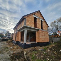 Budorem Developer sp. zo.o. - Surowy stan domu z czerwonej cegły, widoczne betonowe elementy konstrukcyjne, czarna izolacja fundamentów, otoczenie placu budowy i pochmurne niebo.