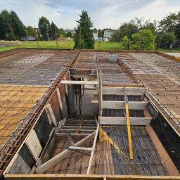 Budorem Developer sp. zo.o. - Zbrojenie stropu z deskowaniem i przygotowanym otworem na schody w domu jednorodzinnym. Widok z góry na plac budowy w słoneczny dzień.
