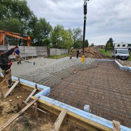 Budorem Developer sp. zo.o. - Wylewanie betonu na zbrojenie fundamentów domu jednorodzinnego. Widoczni pracownicy, pompa do betonu, szalunki z izolacją. W tle koparka i samochód dostawczy.