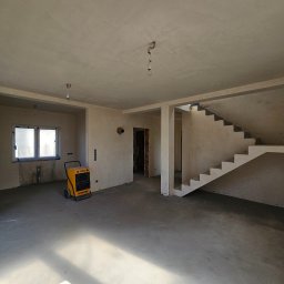 Budorem Developer sp. zo.o. - Surowe wnętrze domu w trakcie budowy: betonowa podłoga, otynkowane ściany, schody w stanie surowym, żółty osuszacz powietrza. Widoczne niedokończone instalacje.