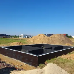 Zabezpieczone, pomalowane na czarno, fundamenty domu jednorodzinnego z widocznymi ścianami fundamentowymi i przestrzenią wewnętrzną, na tle hałdy piasku i zabudowy jednorodzinnej w słoneczny dzień.