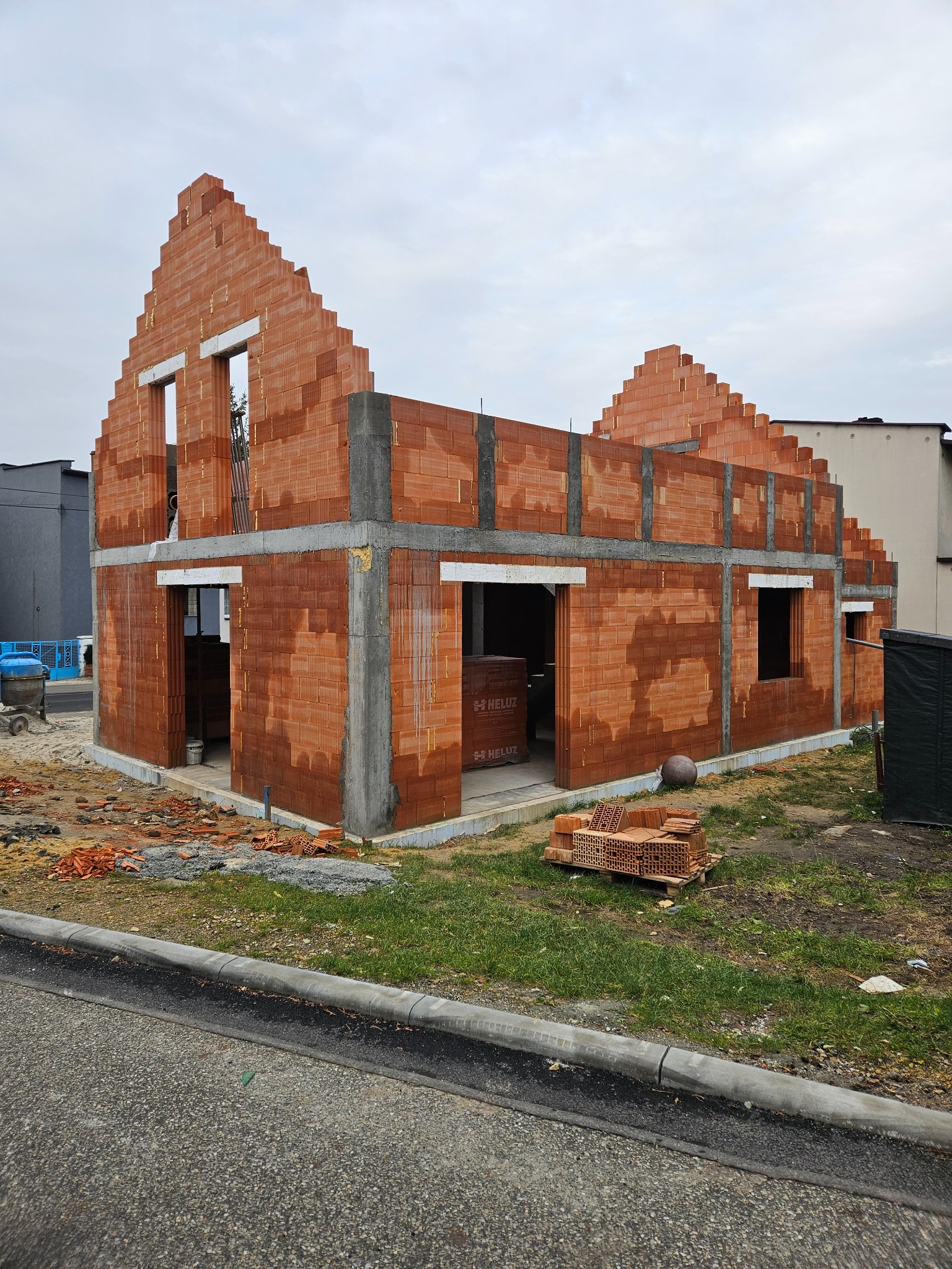Surowy stan domu z czerwonej cegły Heluz, widoczne otwory okienne i drzwiowe, betonowe wzmocnienia, paleta cegieł obok. Budowa w toku, pochmurne niebo w tle.