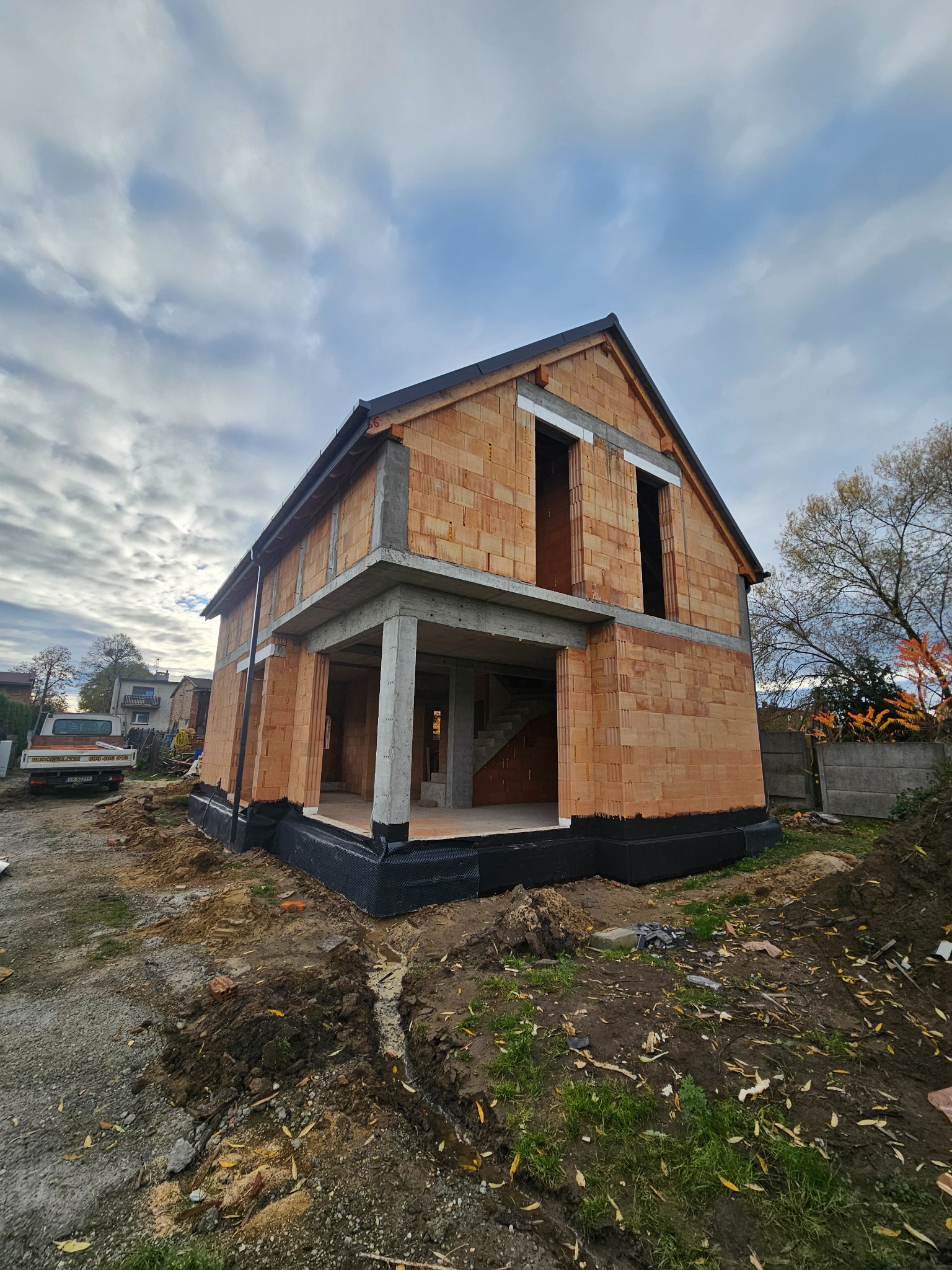 Surowy stan domu z czerwonej cegły, widoczne betonowe elementy konstrukcyjne, czarna izolacja fundamentów, otoczenie placu budowy i pochmurne niebo.