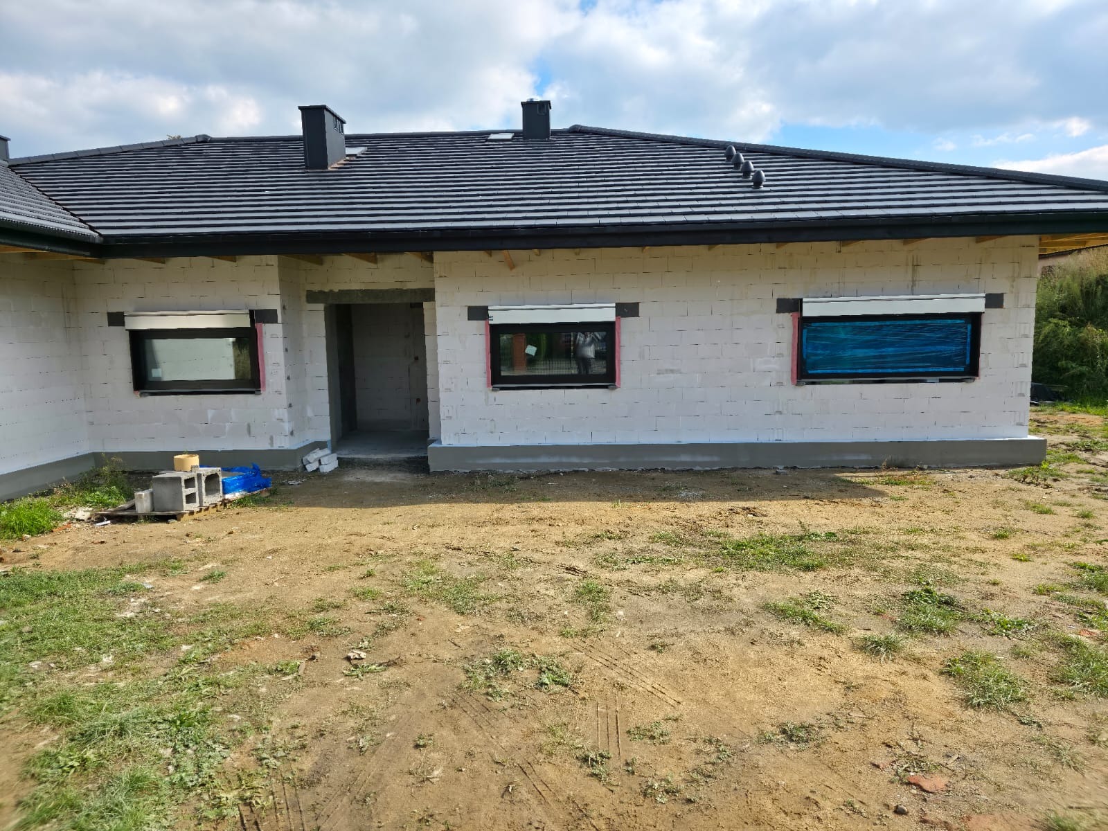Niedokończony dom z białego pustaka z czarną dachówką i oknami z roletami, widok z zewnątrz na trawiastym terenie.