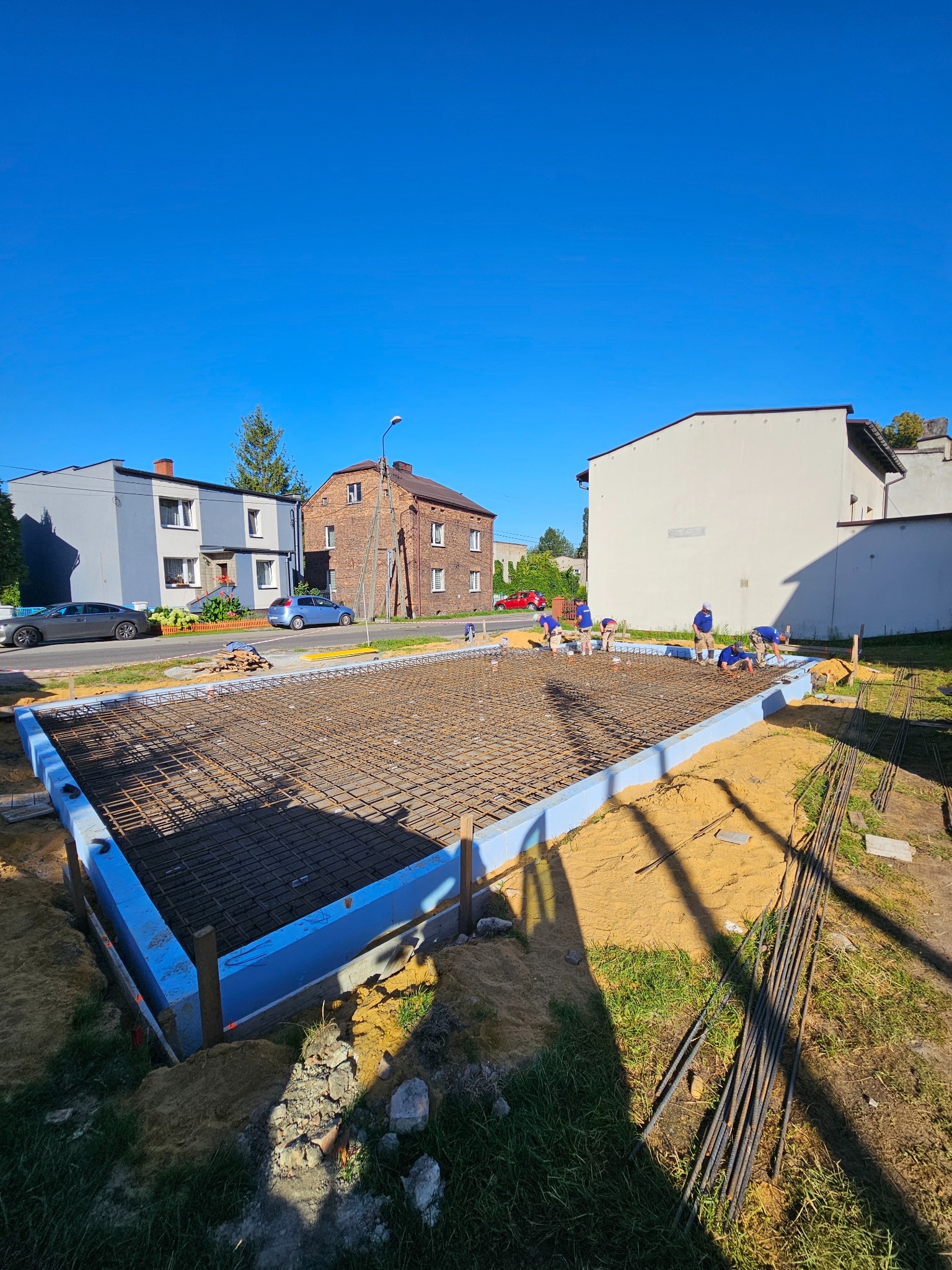 Zbrojenie fundamentów pod budowę domu jednorodzinnego. Widoczni pracownicy w niebieskich koszulkach. Ujęcie w słoneczny dzień, z perspektywy szerokokątnej.