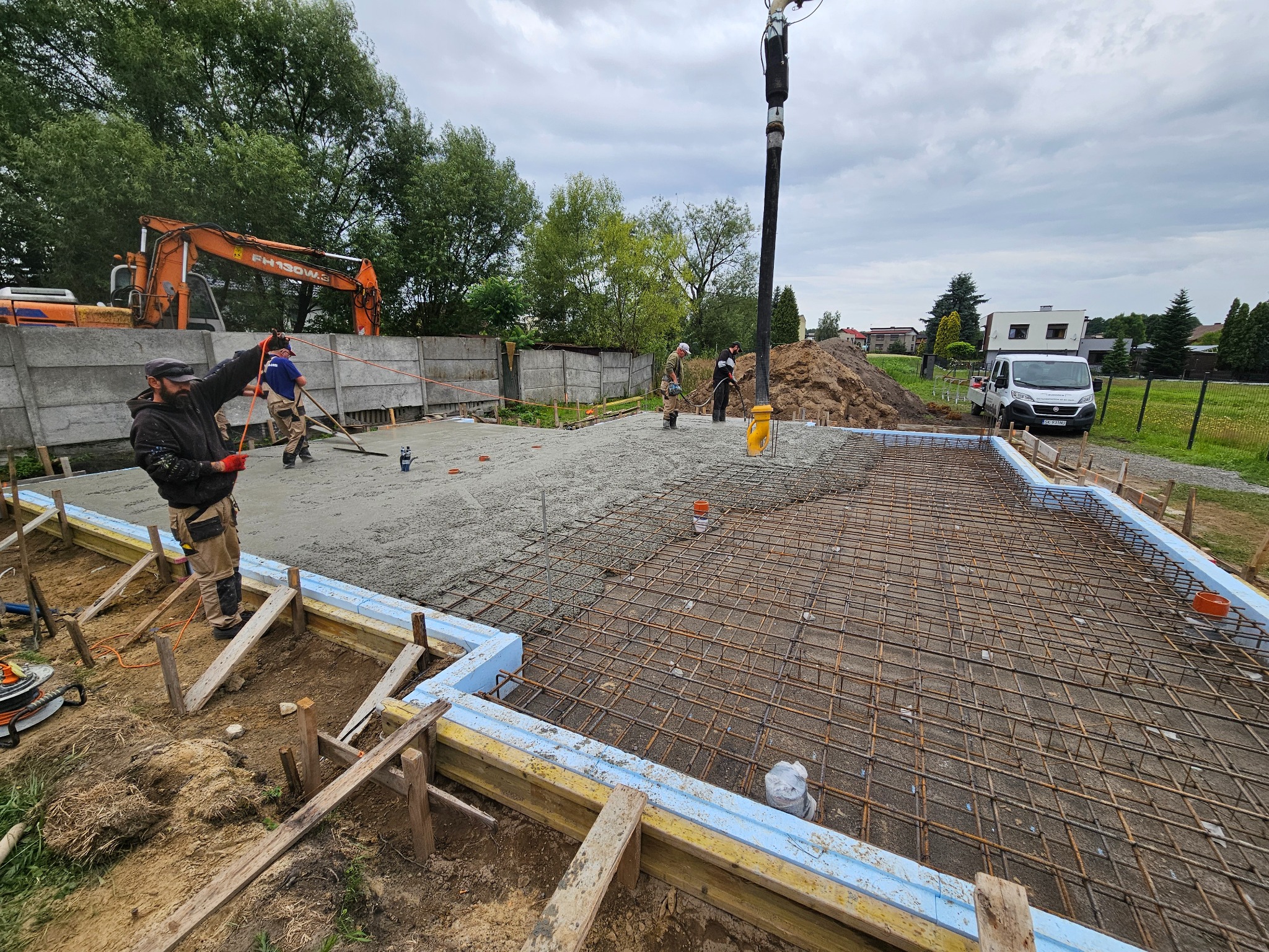 Wylewanie betonu na zbrojenie fundamentów domu jednorodzinnego. Widoczni pracownicy, pompa do betonu, szalunki z izolacją. W tle koparka i samochód dostawczy.
