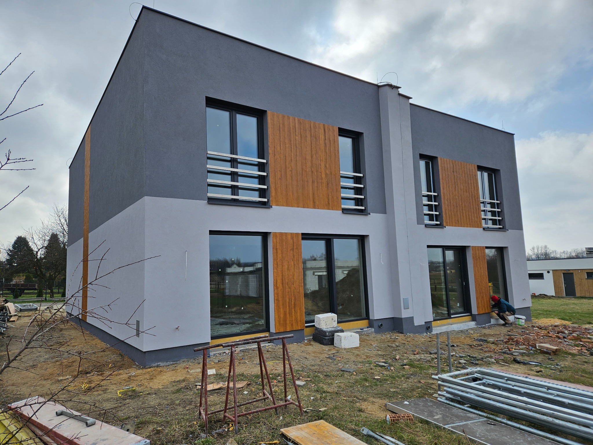 Dwukondygnacyjny dom z szarą elewacją i drewnianymi panelami między oknami. Na pierwszym planie rusztowania i materiały budowlane, sugerujące prace wykończeniowe.
