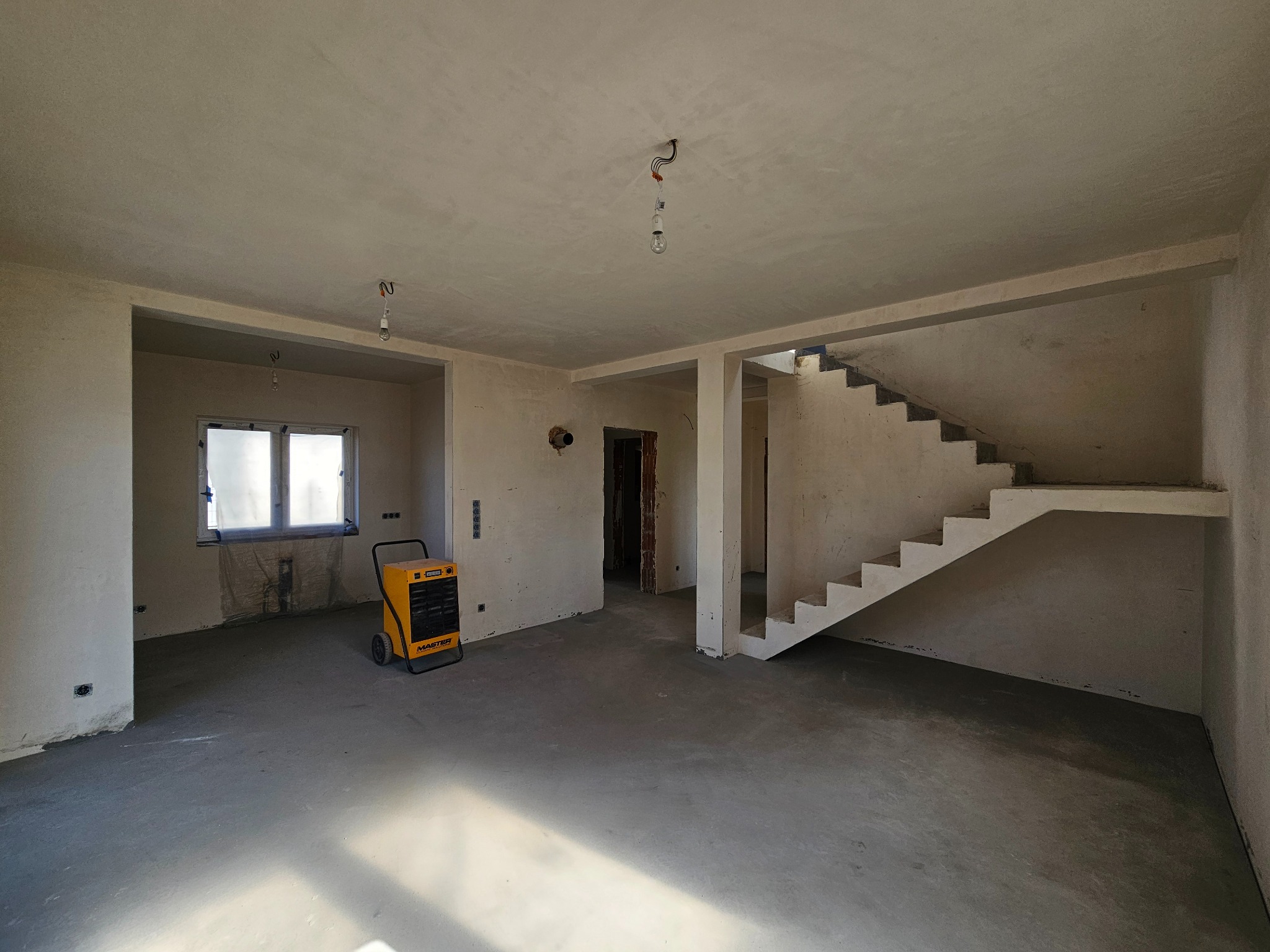 Surowe wnętrze domu w trakcie budowy: betonowa podłoga, otynkowane ściany, schody w stanie surowym, żółty osuszacz powietrza. Widoczne niedokończone instalacje.