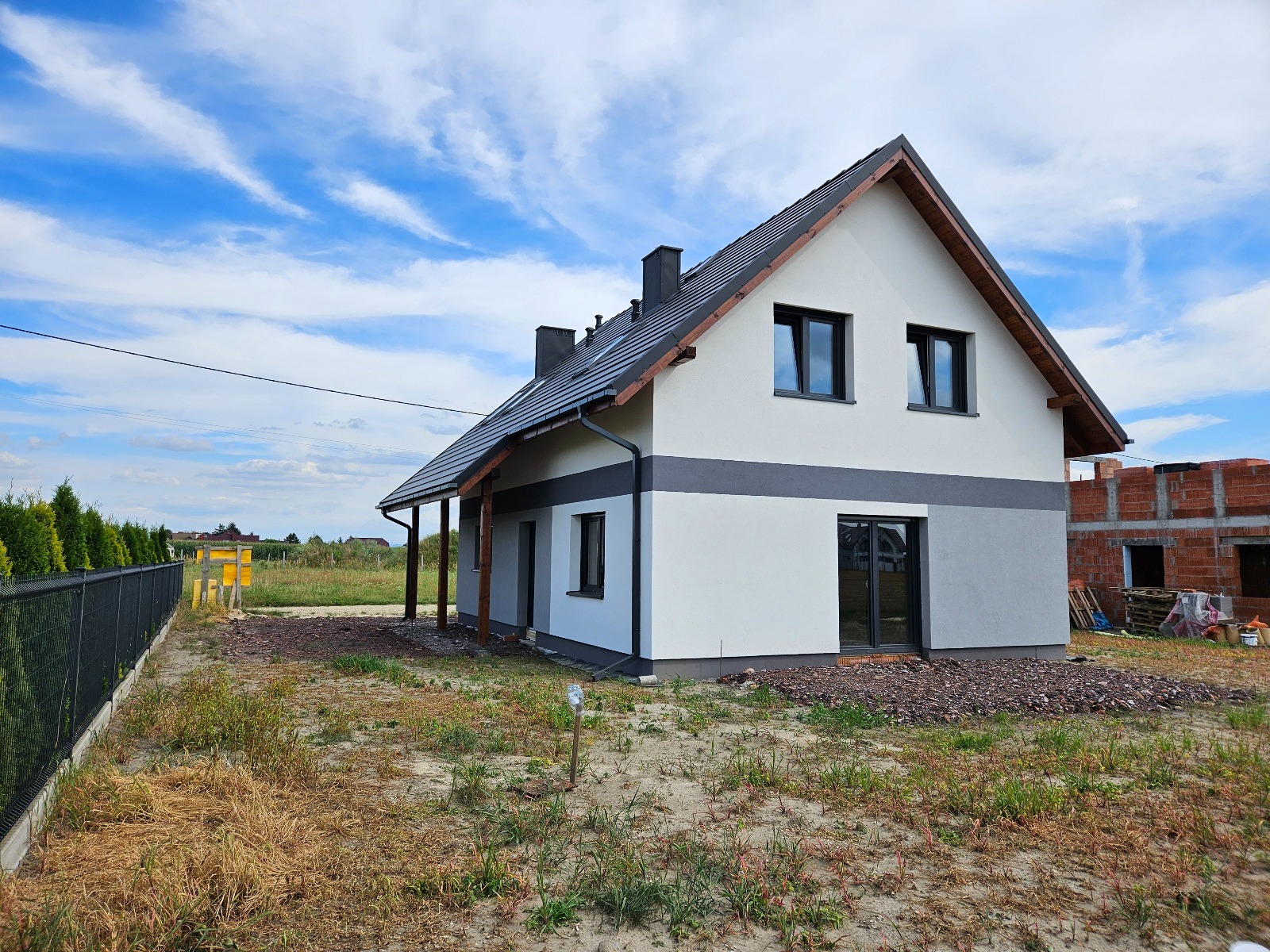 Nowoczesny dom jednorodzinny z szarą elewacją i ciemną stolarką okienną, widoczny z boku, z fragmentem budowy w tle i niezagospodarowanym terenem.