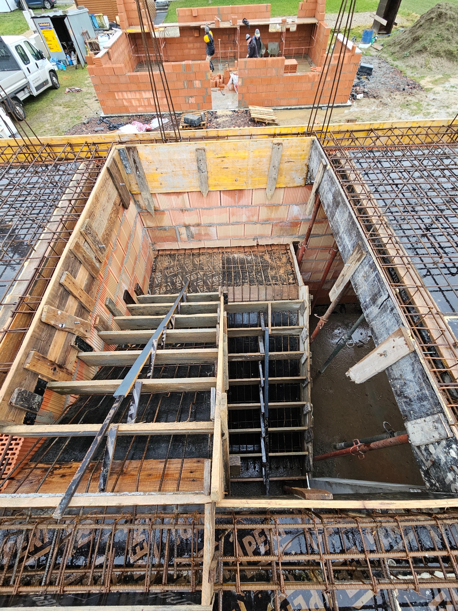 Z góry widok na szalunek schodów zbrojonych stalą, w tle fragment murowanej konstrukcji budynku z czerwonej cegły. Widoczne drewniane elementy konstrukcyjne i stalowe pręty zbrojeniowe.